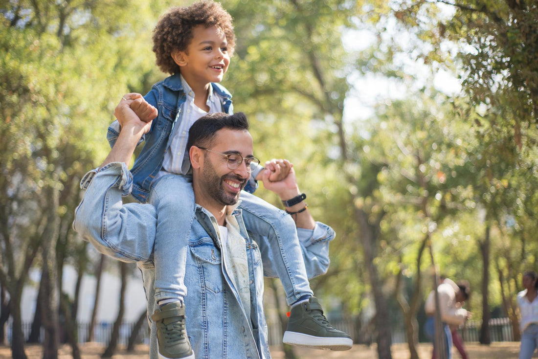 Smiling father carrying his young son on his shoulders during a sunny walk in the park, representing joyful Father’s Day moments and thoughtful gift ideas.