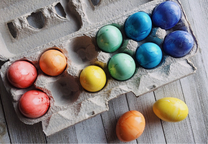 Naturally dyed Easter eggs in vibrant shades of pink, orange, yellow, green, blue, and purple, displayed in a recycled egg carton on a wooden surface