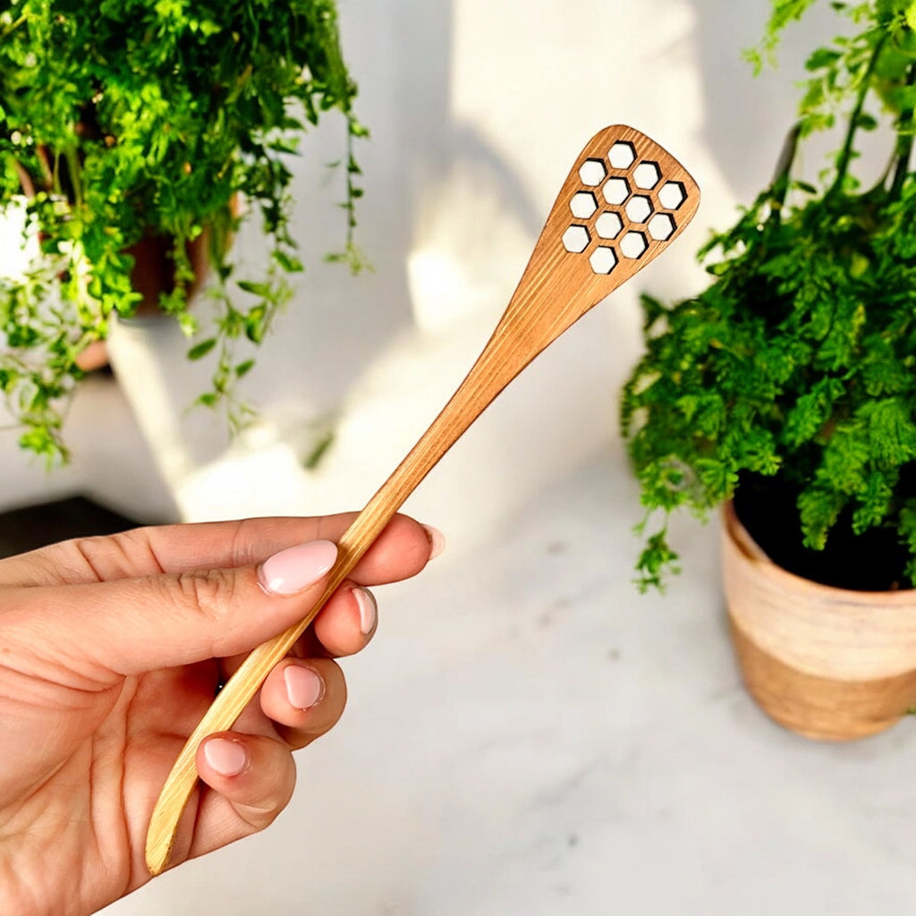 Hand holding a bamboo honey dipper with a honeycomb pattern against a blurred indoor background with plants.
