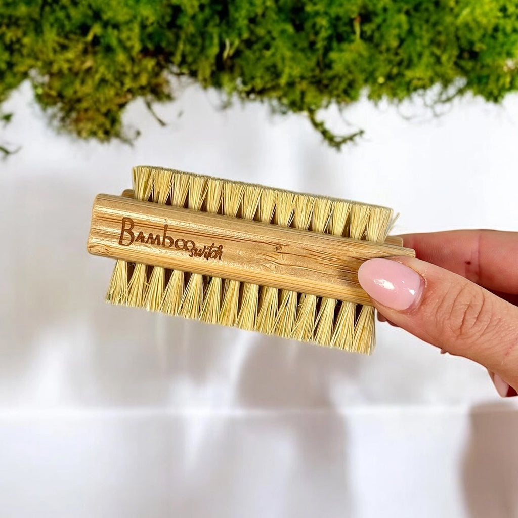 Hand holding a bamboo nail brush against a blurred natural background.