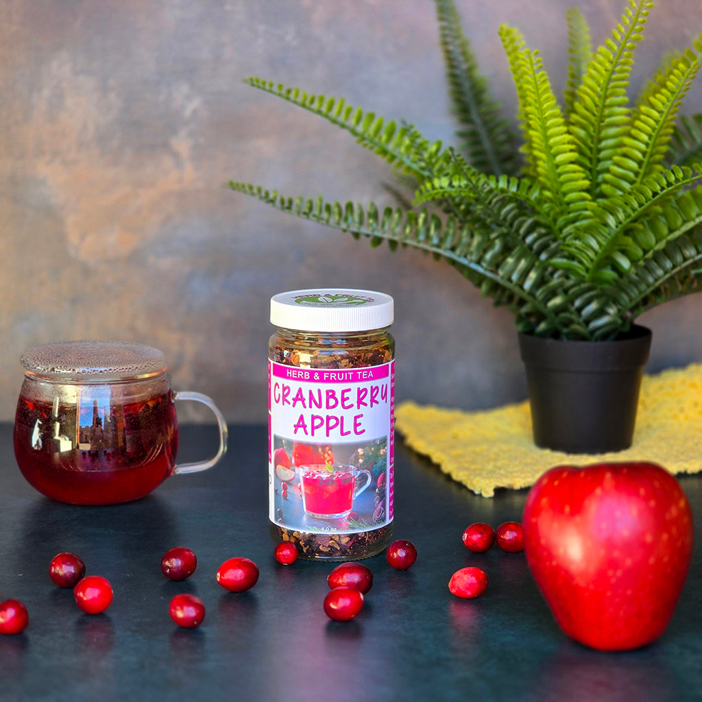 Jar of Cranberry Apple Herb & Fruit Tea on a dark surface surrounded by cranberries, an apple and a cup of tea brewing.