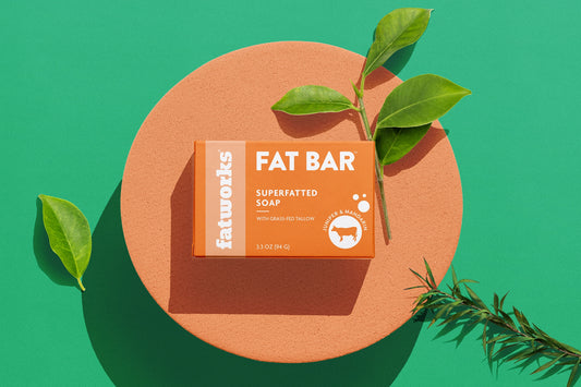 A bar of Fat Bar Superfatted Tallow Soap by Fatworks placed on a circular orange platform with green leaves around it.