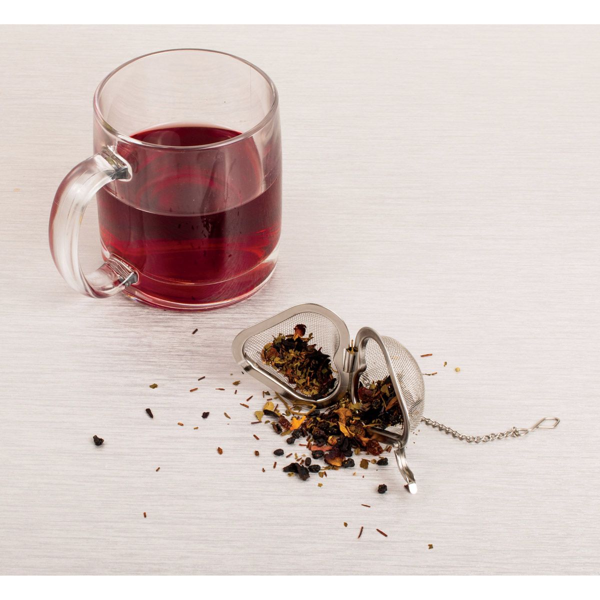 Clear mug with red tea and stainless steel heart tea infuser on a white surface