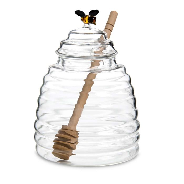 Clear glass beehive-shaped jar with wooden dipper and decorative bee on a white background