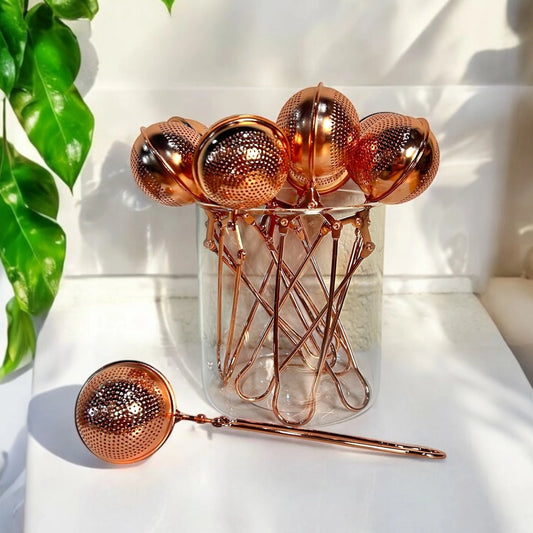 Set of rose gold tea infusers with a clear jar on a white surface.