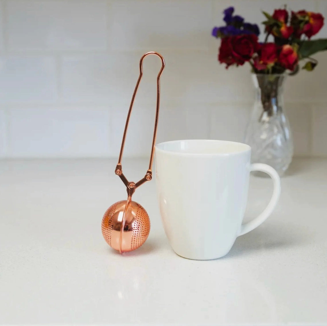 Rose gold tea ball infuser next to a white mug on a light surface with flowers in the background.
