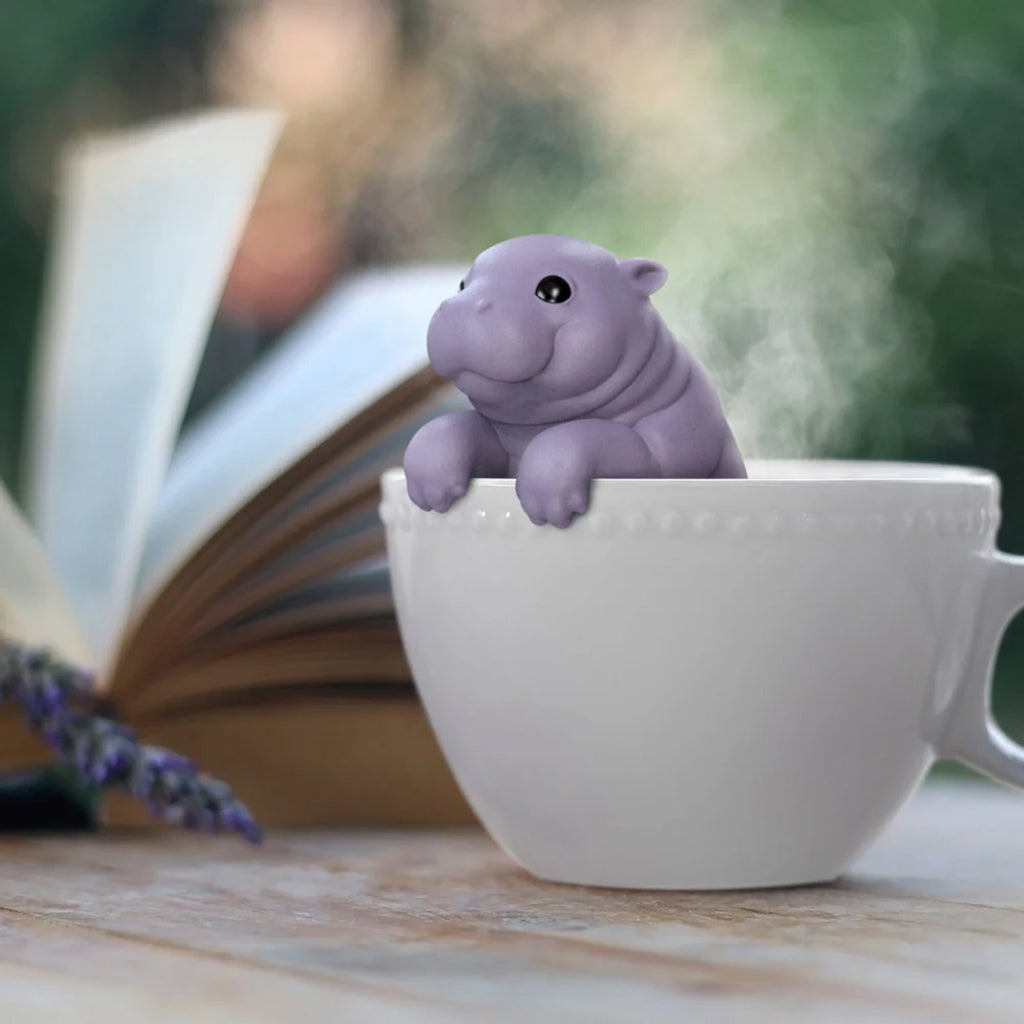 Hippo shaped tea infuser in a white cup with a book and lavender spring in the background