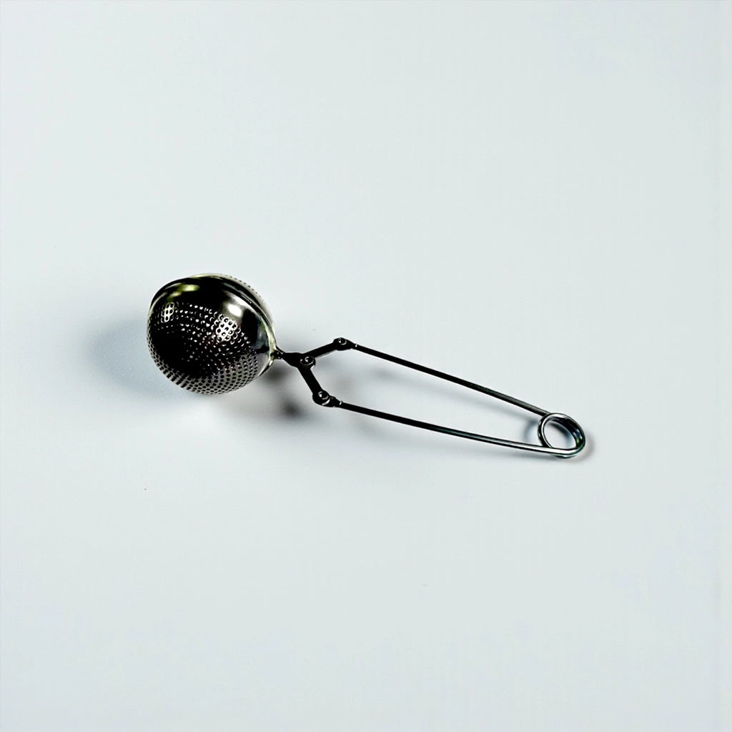 A black metal tea infuser with a perforated spherical mesh head and long spring handles, shown on a plain light gray background.