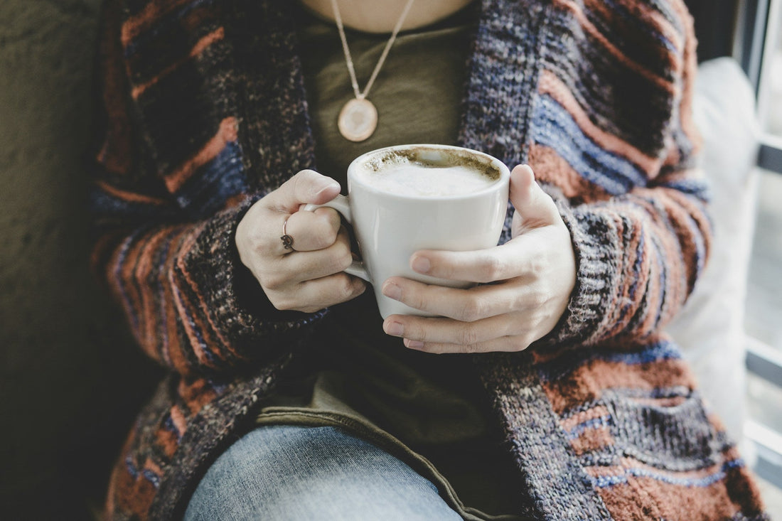Woman wearing a cozy autumn sweater holding a warm mug of coffee or tea near a window.