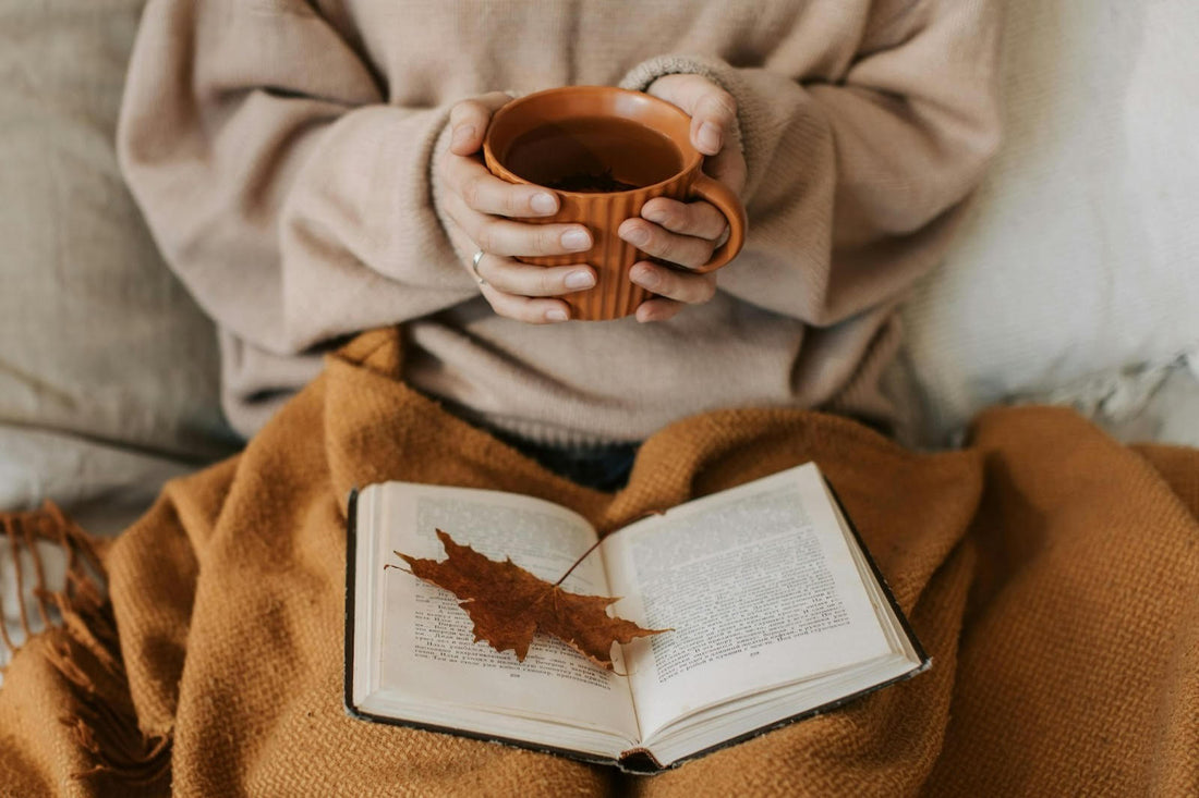 A woman holding a cup of tea with a sweater, blanket and a book with autumn leaf.