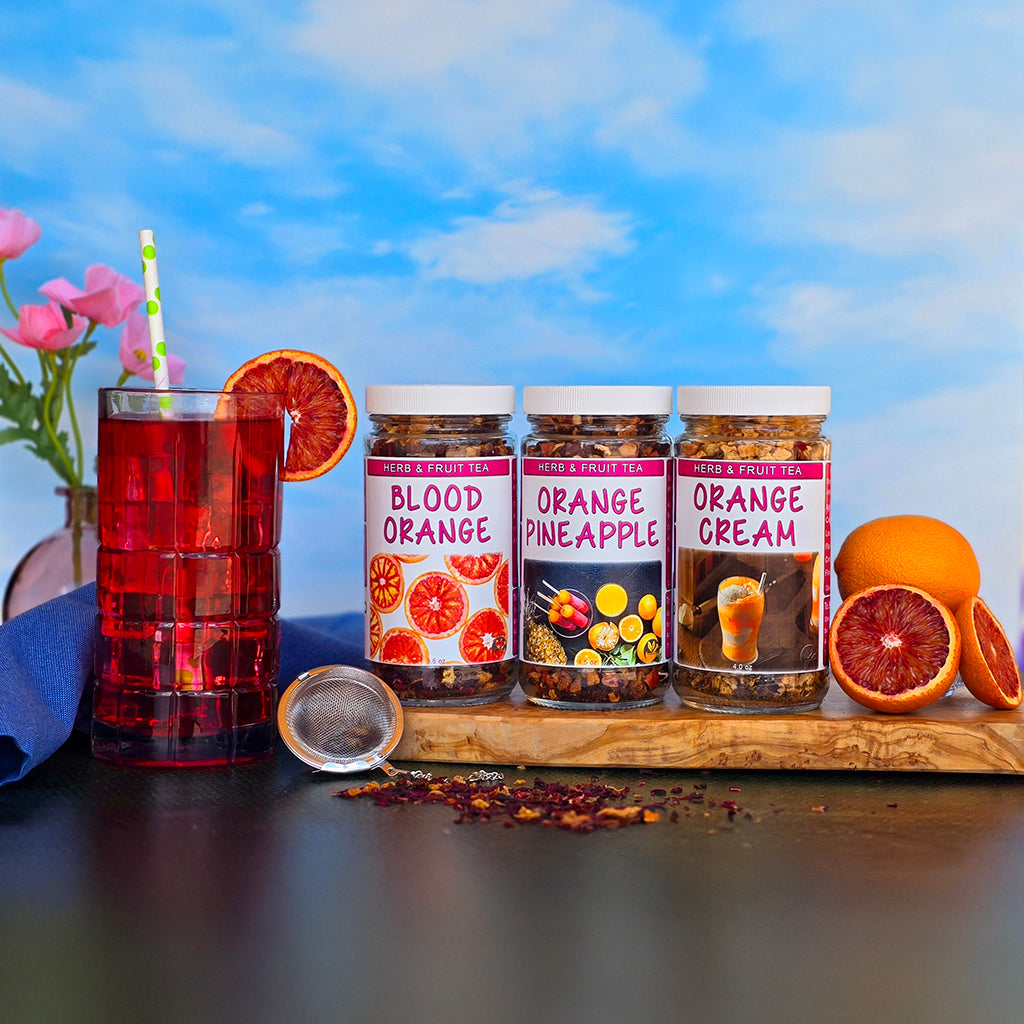 Blood Orange, Orange Pineapple, and Orange Cream loose-leaf teas in glass jars with a tall iced tea, fresh oranges, and a tea infuser from Herb Stop’s Summer Tea Collection