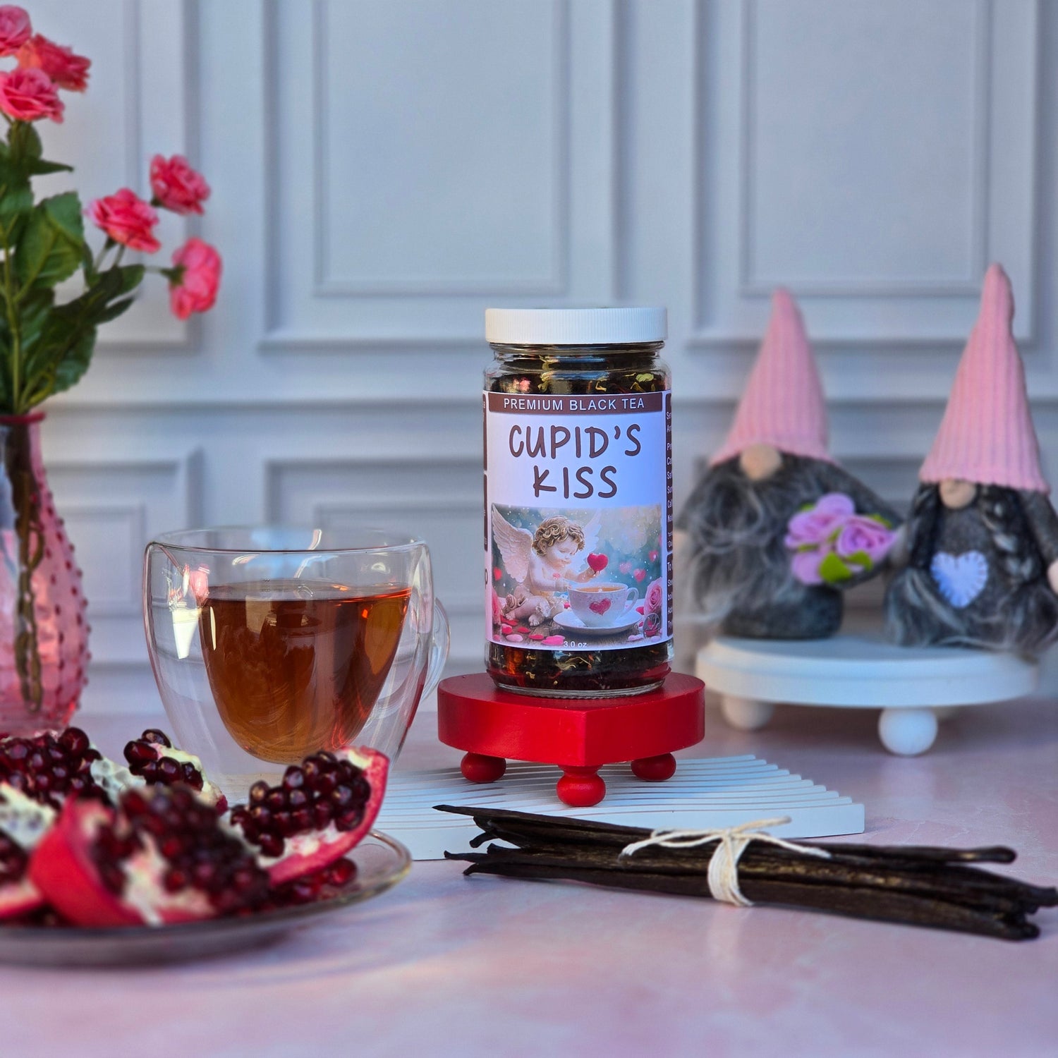 Cupid’s Kiss Black Tea in a glass jar on a red stand, with a clear glass mug of brewed tea, fresh pomegranate, vanilla beans, pink roses, and Valentine gnome decorations from Herb Stop’s Valentine’s Tea Collection