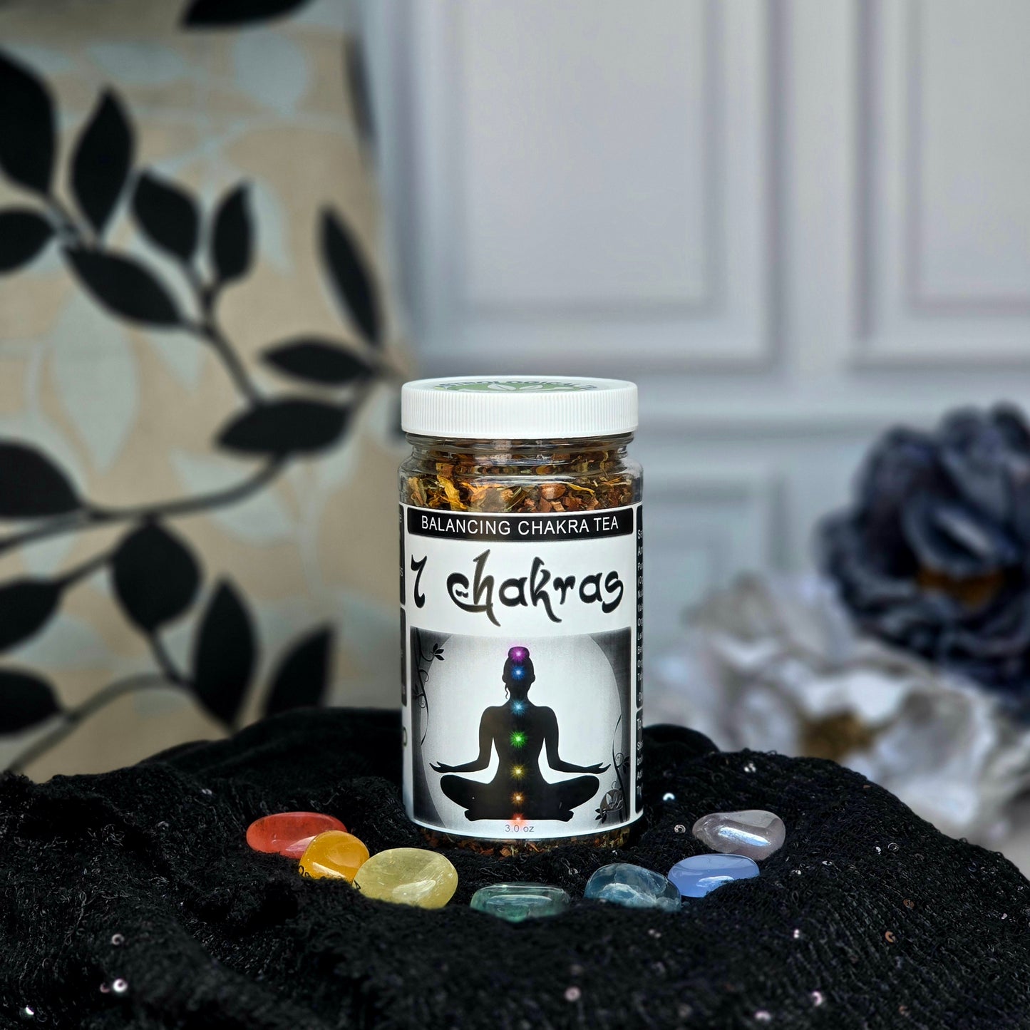 7 Chakras Balancing Herbal Tea jar surrounded by 7 chakra stones on a black cloth with flowers in the background