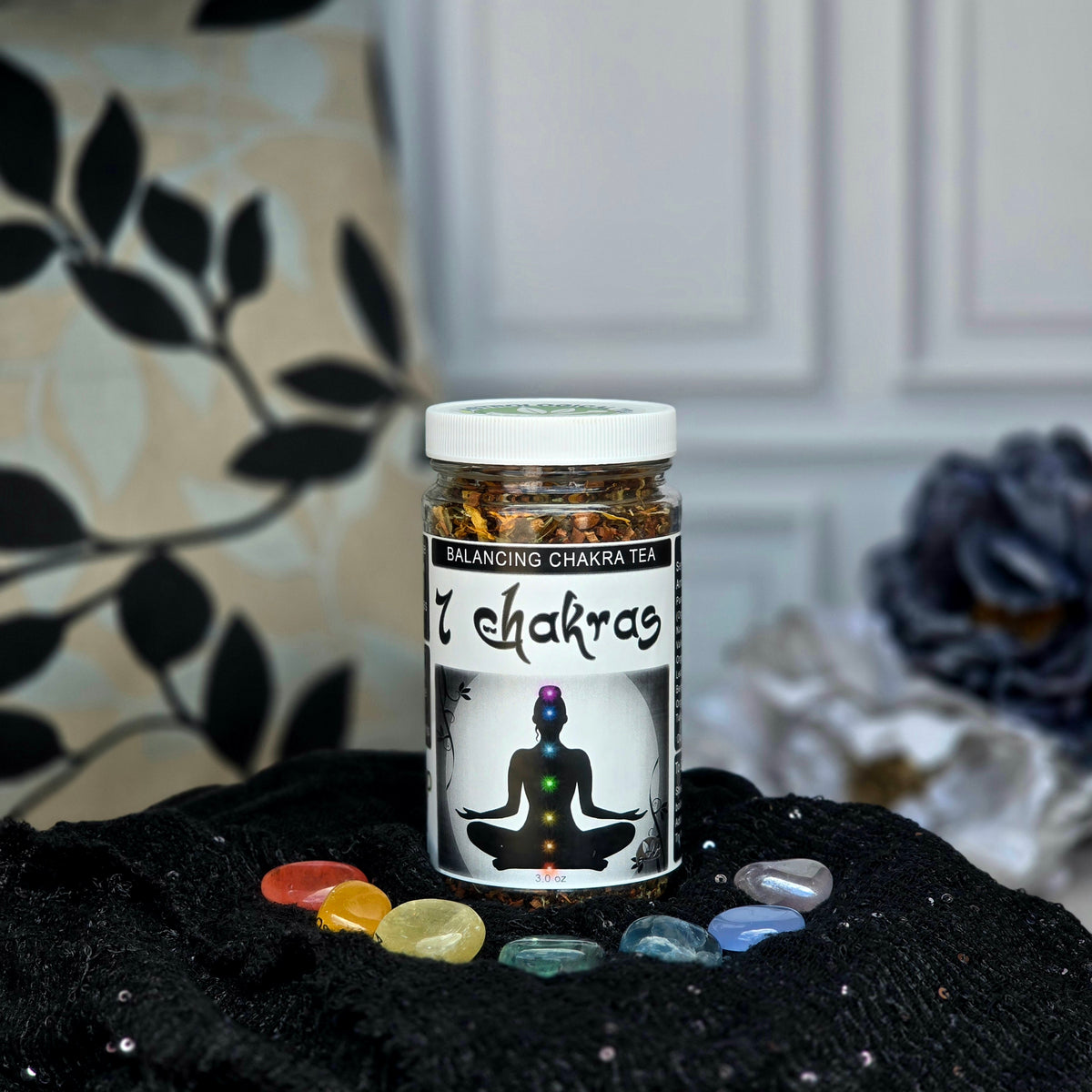 7 Chakras Balancing Herbal Tea jar surrounded by 7 chakra stones on a black cloth with flowers in the background