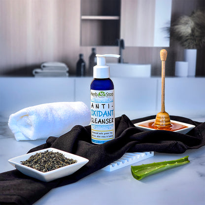 Bottle of Herb Stop Antioxidant Cleanser on a bathroom counter with honey, tea leaves, and aloe vera.
