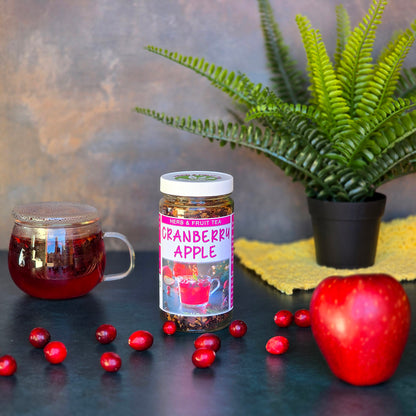 Jar of Cranberry Apple Herb & Fruit Tea on a dark surface surrounded by cranberries, an apple and a cup of tea brewing.