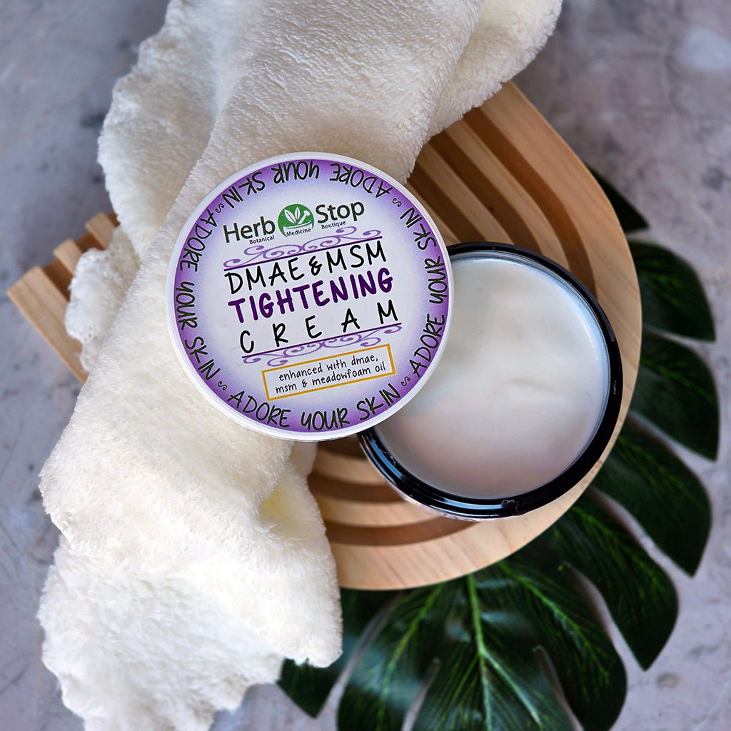 Jar of DMAE & MSM Tightening Cream with a towel and green leaves on a wooden stand.