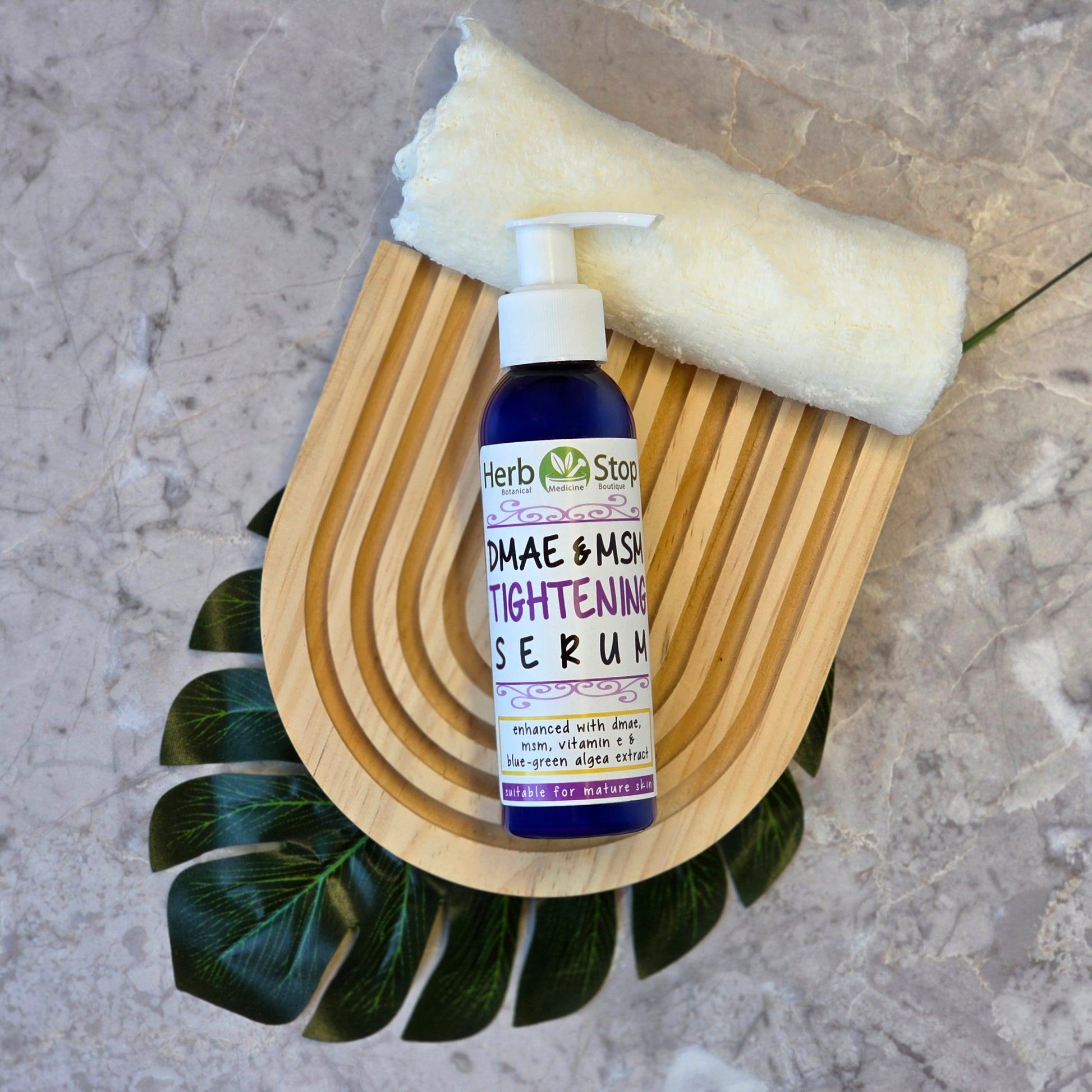 Herb Stop DMAE & MSM Tightening Serum bottle on a wooden soap dish with a leaf and towel in the background.