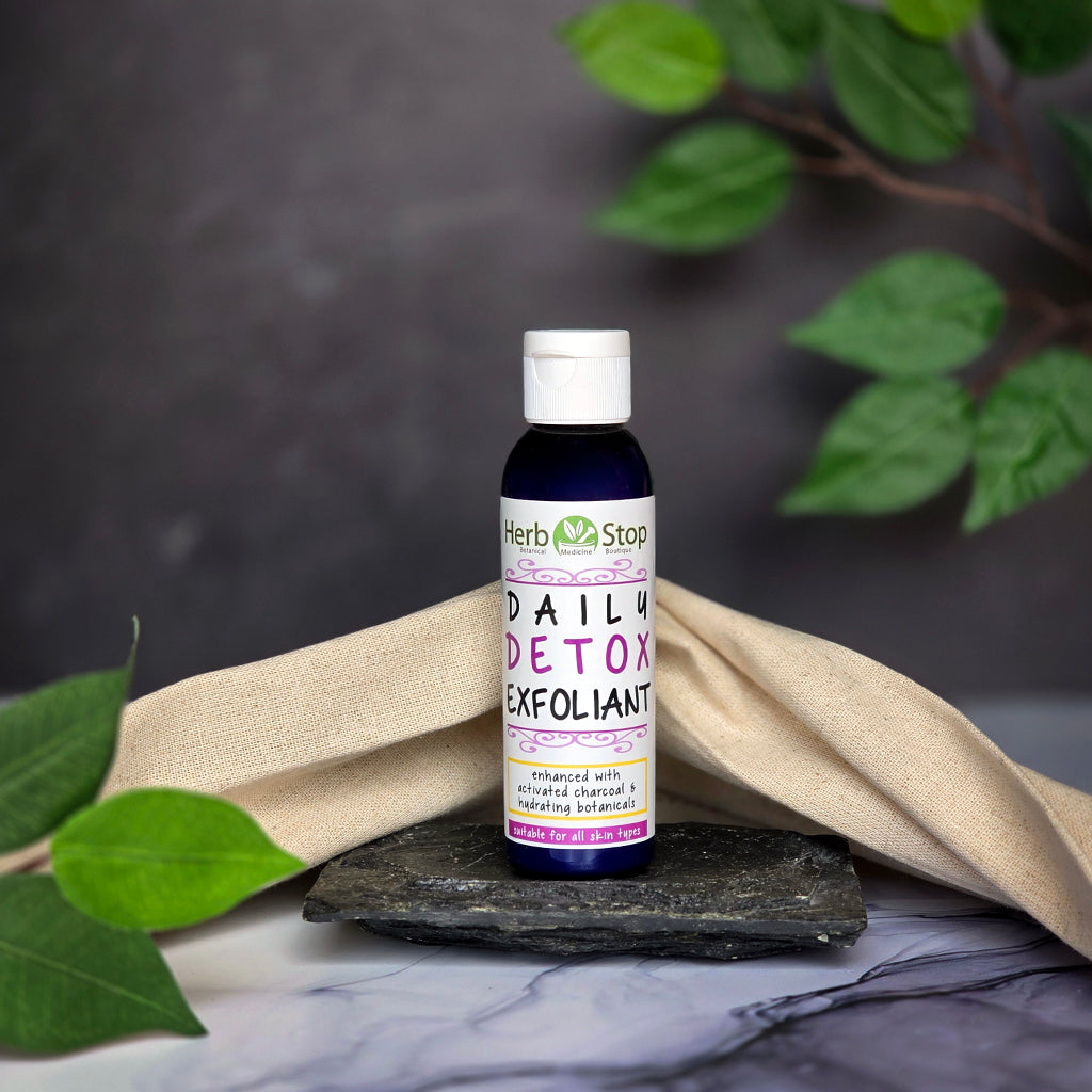 Bottle of Herb Stop Daily Detox Exfoliant on a dark surface with green leaves in the background