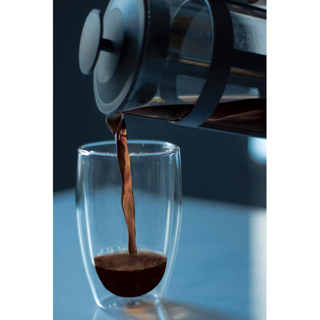Coffee being poured from a glass french press into a clear glass on a reflective surface.
