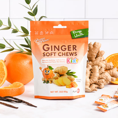 A package of Prince of Peace Kids Ginger Soft Chews in Orange Vanilla flavor, styled with fresh ginger roots, sliced oranges, vanilla beans, and green leaves on a white tiled background.