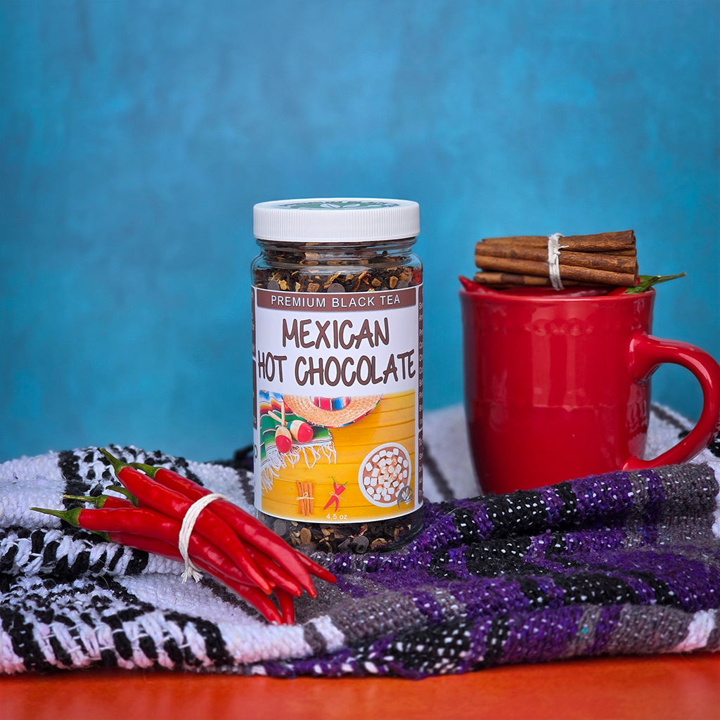 A jar of Mexican Hot Chocolate premium black tea stands on a patterned cloth next to a red mug with cinnamon sticks and red chili peppers, set against a textured blue background.