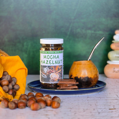 A jar of Mocha Hazelnut Yerba Mate tea on a plate with chocolate, hazelnuts and a gourd with a bombilla . 