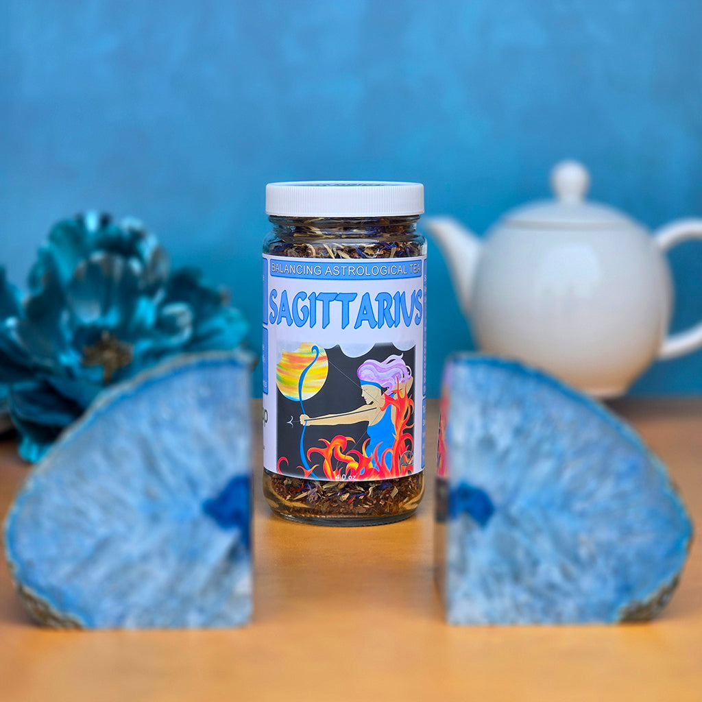 Sagittarius Tea on a sand surface with a blue background, teapot in the back and blue agate crystals in the foreground.