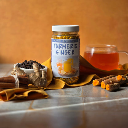 Jar of Herb Stop Turmeric Ginger organic herbal tea with a cup of tea in the background and surrounded by turmeric, ginger, black pepper and cinnamon. 