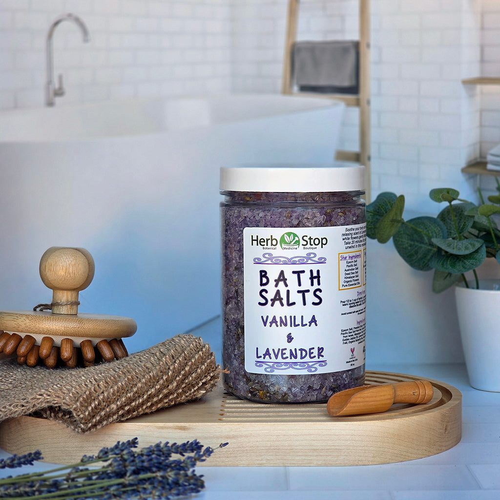 A jar of Herb Stop Vanilla & Lavender bath salts sits on a wooden tray in a bathroom setting with a bathtub, wooden scoop, burlap cloth, and lavender sprigs.