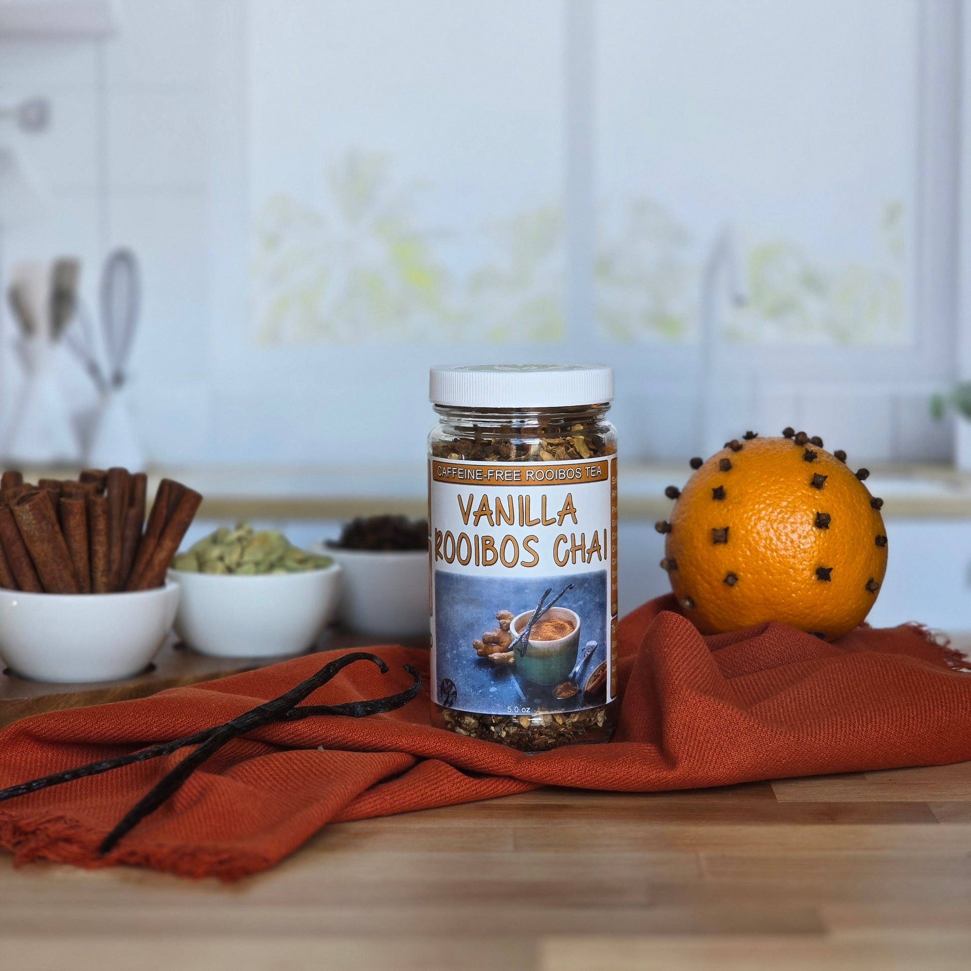 A jar of Vanilla Rooibos Chai surrounded by cinnamon, cardamom and an orange on a wood surface