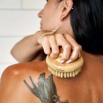 Woman using a bamboo brush on their back with a white tiled wall in the background