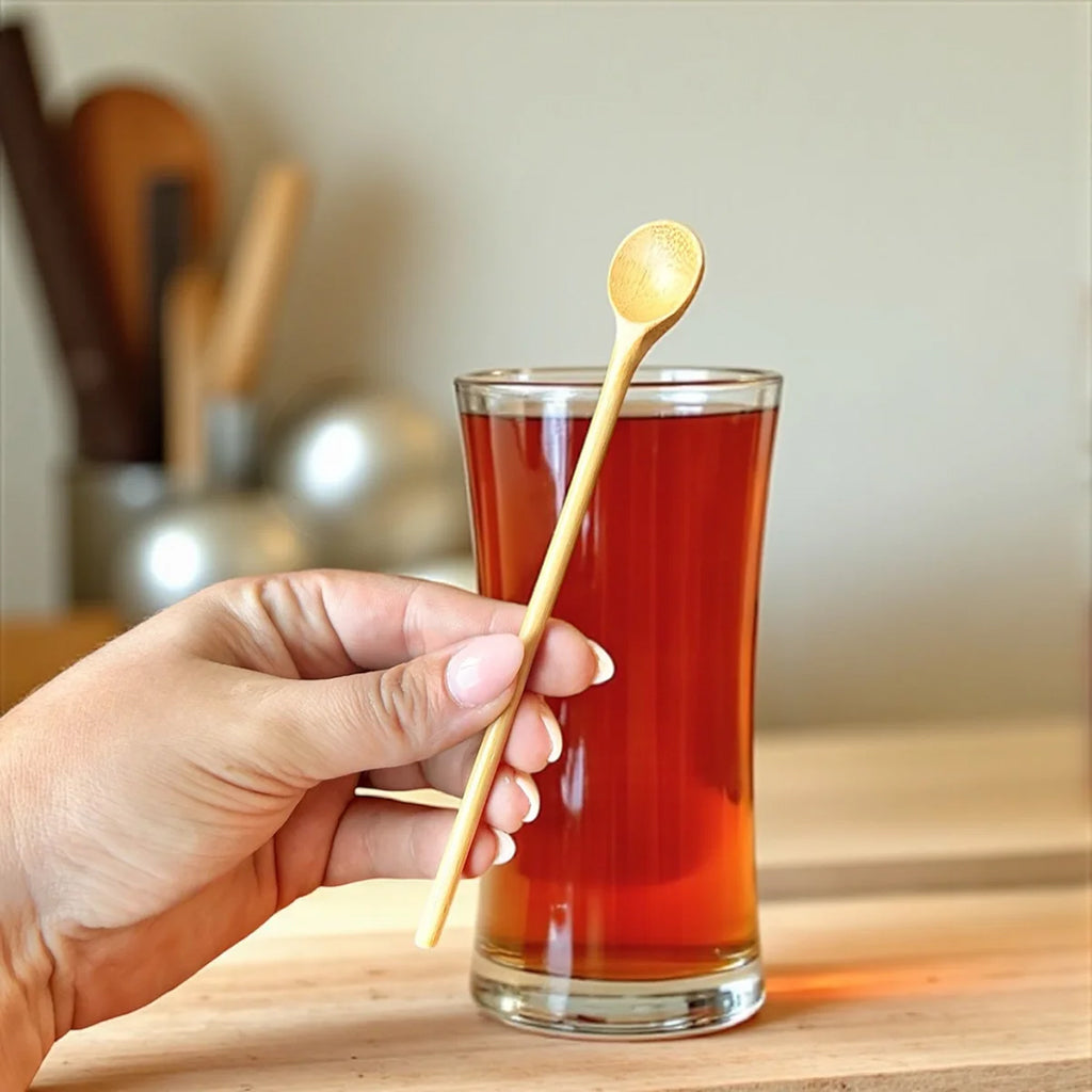 Hand holding a bamboo spoon near a glass of tea with a blurred kitchen background