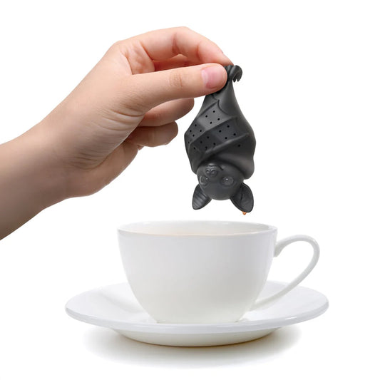 A hand holds a black bat-shaped tea infuser above a white ceramic teacup and saucer on a white background.