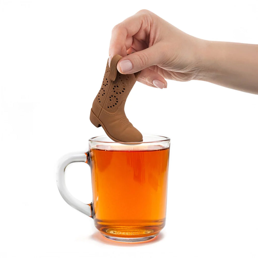 A hand holds a small cowboy boot-shaped tea infuser above a clear glass mug filled with tea on a white background.