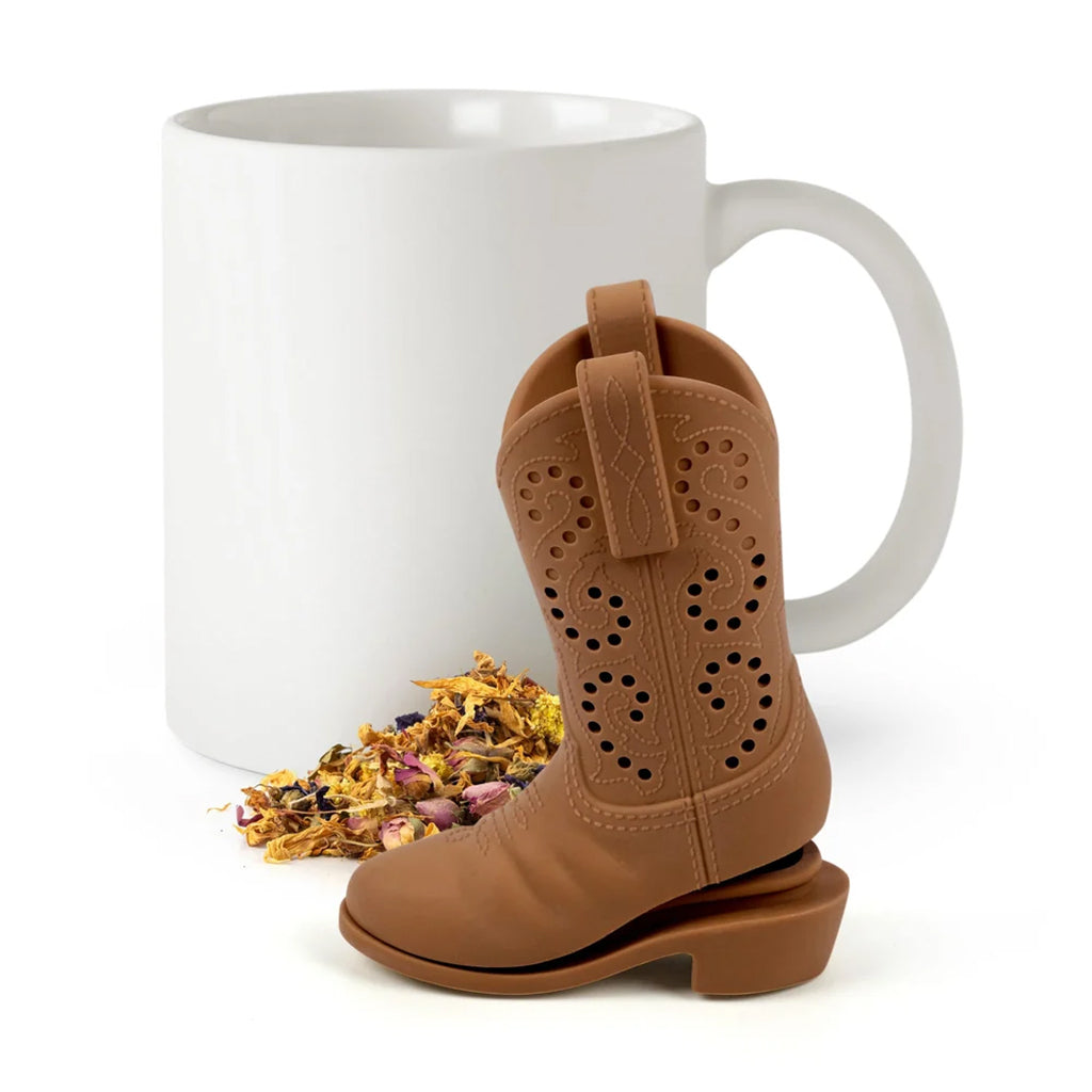 A miniature brown cowboy boot tea infuser stands in front of a white ceramic mug on a white background, with dried tea and flower petals at their base.