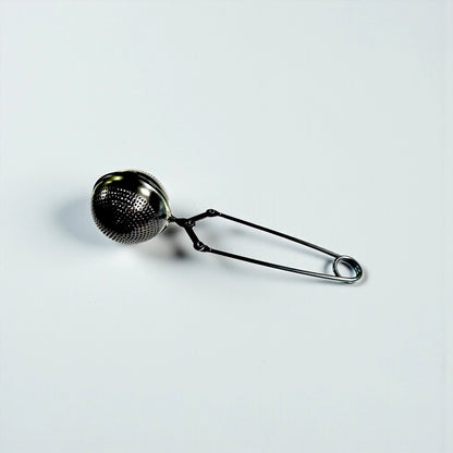 A black metal tea infuser with a perforated spherical mesh head and long spring handles, shown on a plain light gray background.