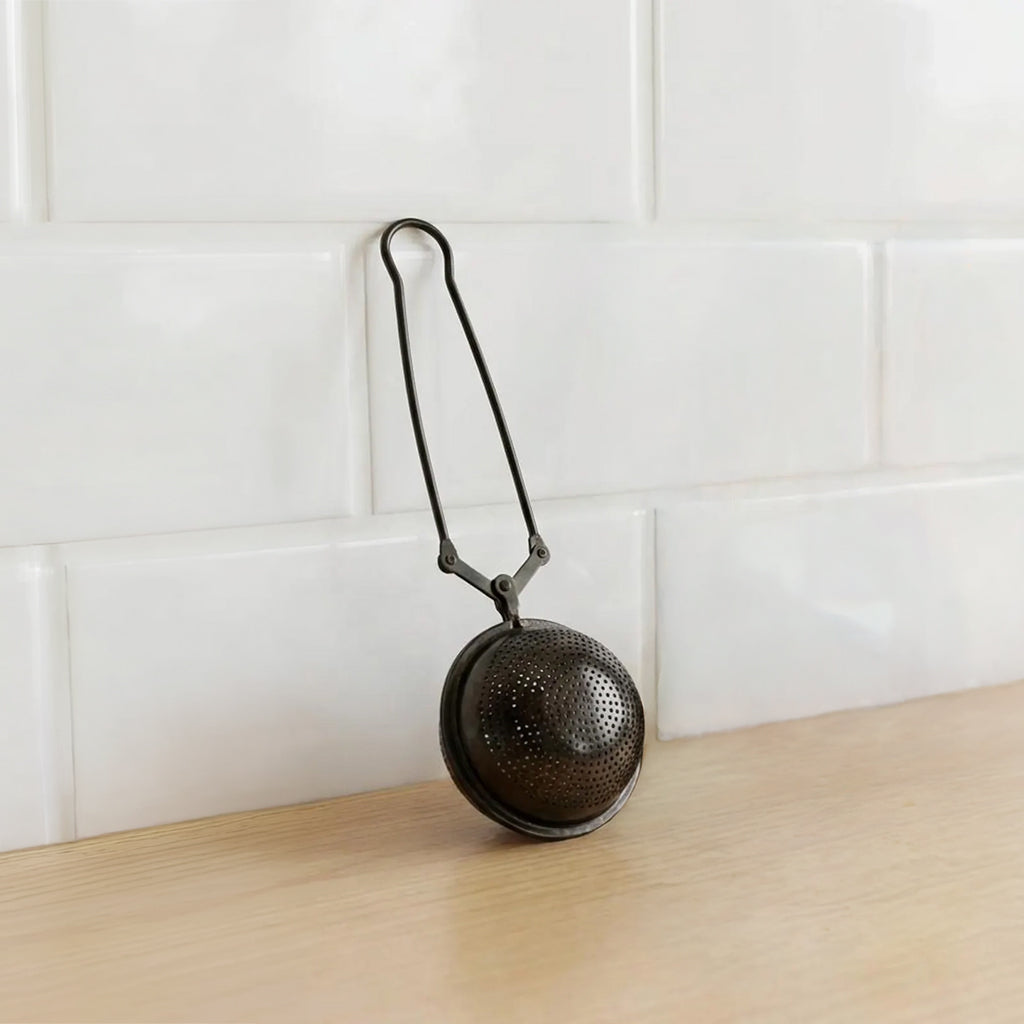 A round metal tea tea infuser with a hanging loop, resting on a light wood surface against a white subway tile backsplash.