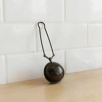 A round metal tea tea infuser with a hanging loop, resting on a light wood surface against a white subway tile backsplash.