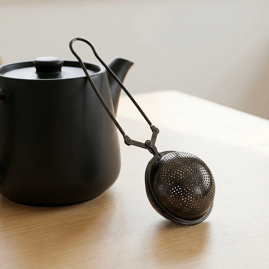 Black tea ball infuser with a black teapot on a wooden surface