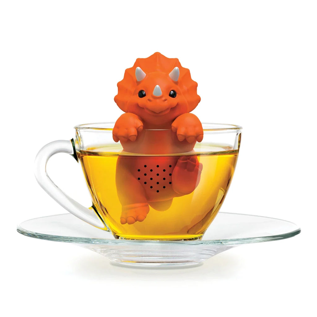 An orange dinosaur-shaped tea infuser sits inside a clear glass teacup filled with tea on a matching glass saucer, photographed against a white background.